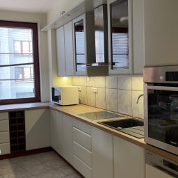 Vlad Tracenko - Jasna kuchnia z białymi szafkami, drewnianym blatem, zabudowaną kuchenką i zlewem, widok na okno z roletami i zabudowę na wino.