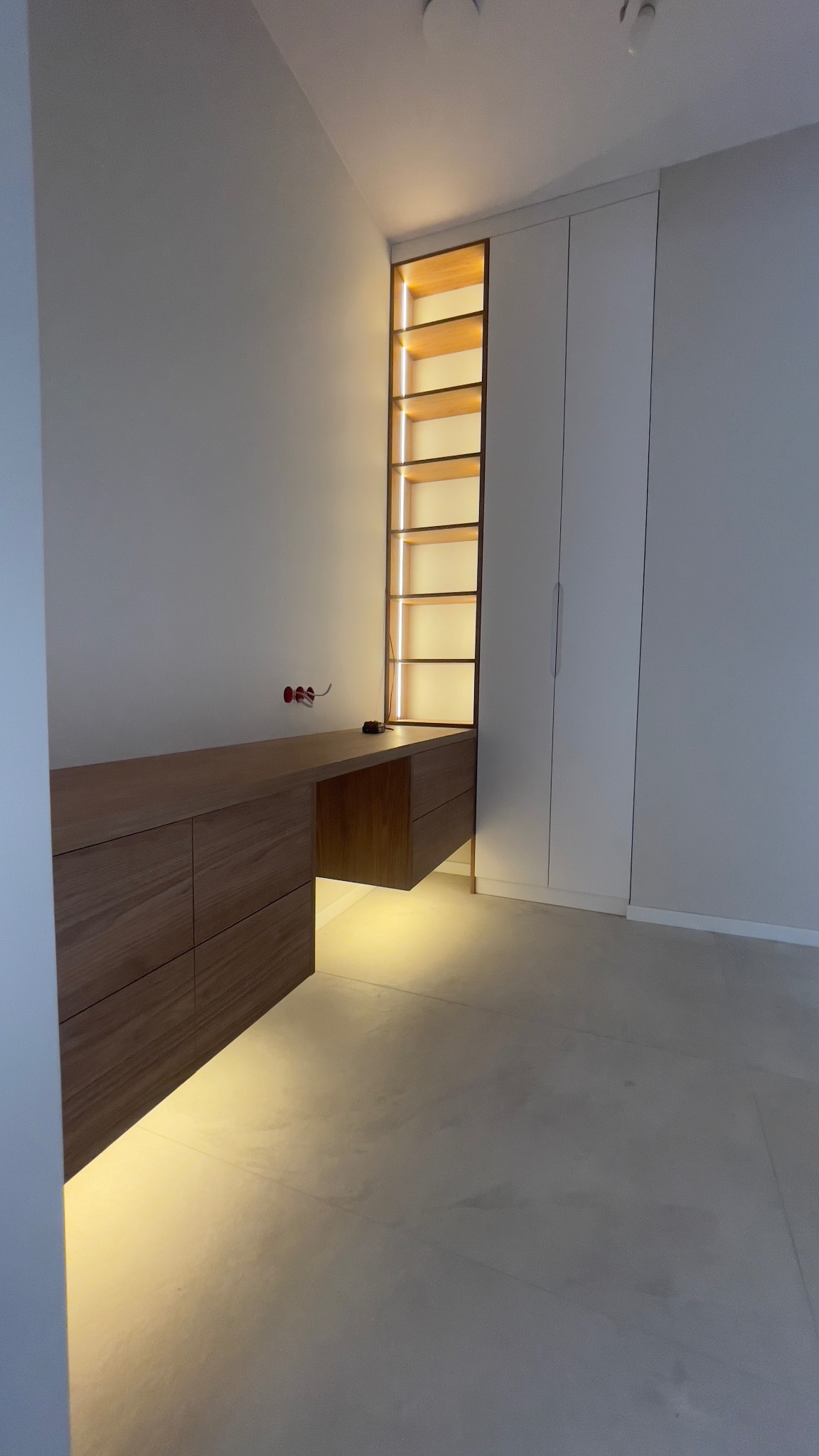 Drewniane biurko z szufladami i otwartą przestrzenią, wbudowane w ścianę obok białej szafy, z oświetleniem LED podkreślającym półki i przestrzeń pod biurkiem, widoczne gniazdka elektryczne na ścianie.