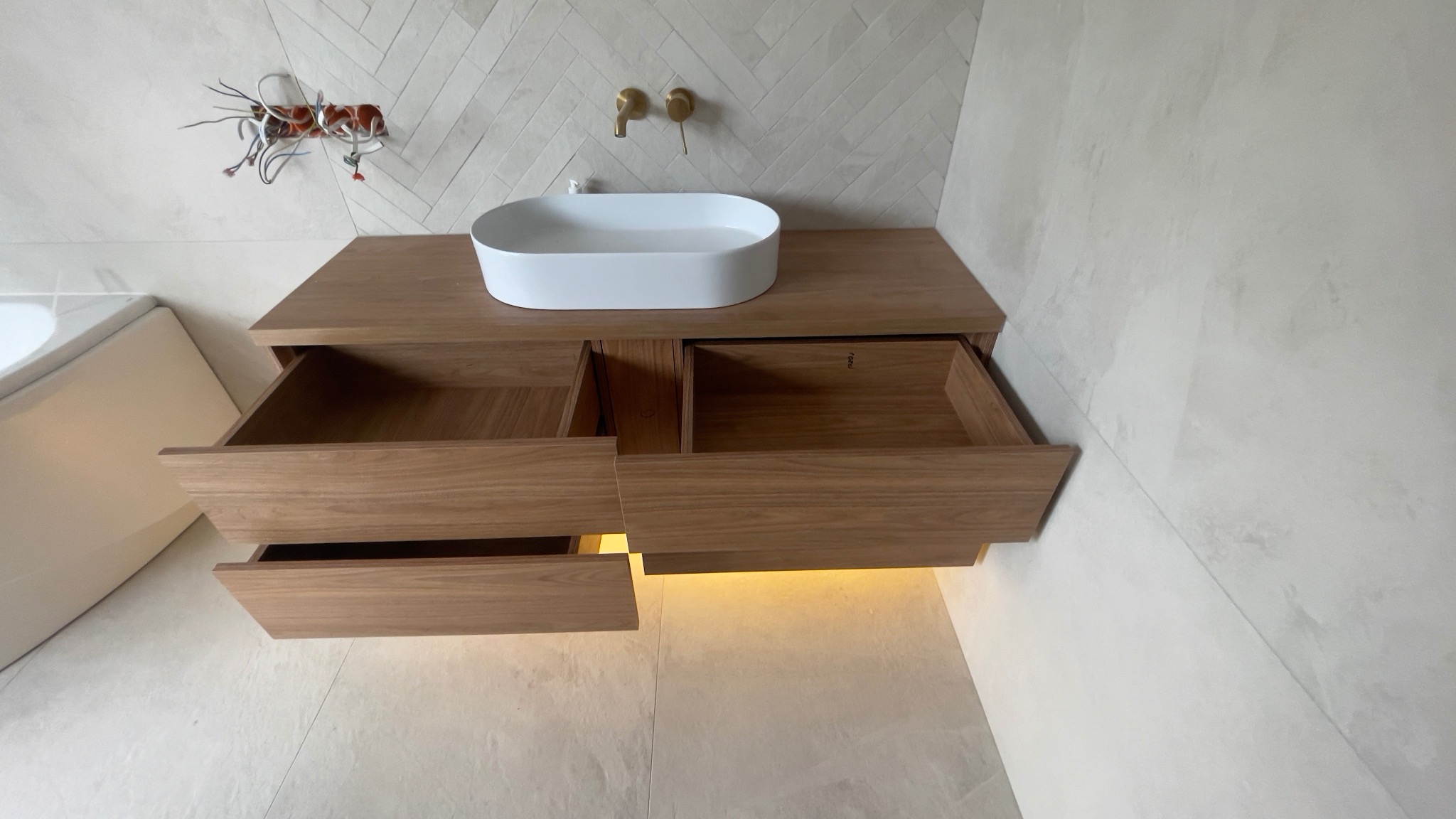 Drewniana szafka pod umywalkę z dwoma wysuniętymi szufladami i podświetleniem LED, umywalka nablatowa, bateria umywalkowa w kolorze złotym, widoczne kable instalacji elektrycznej.