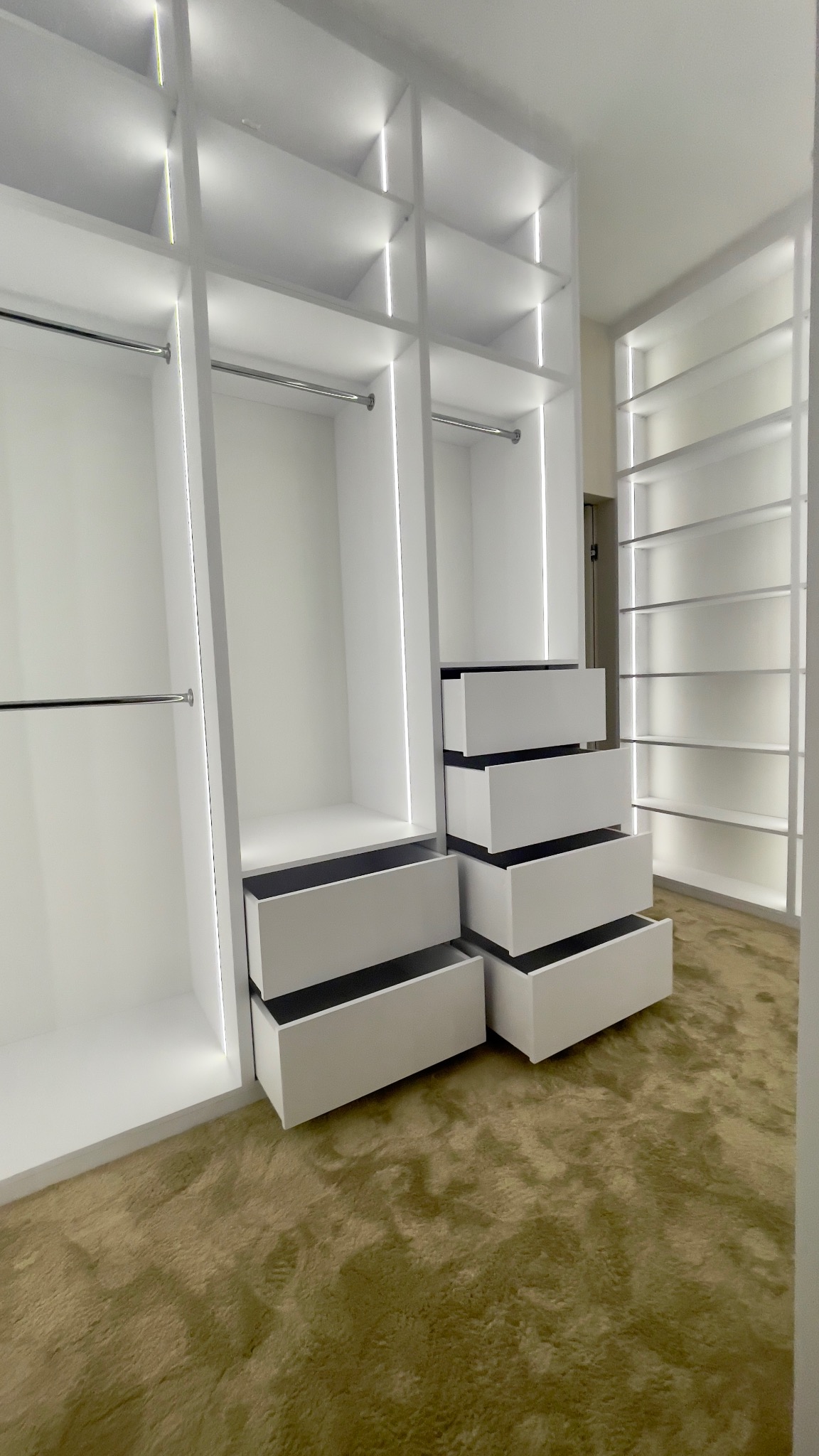 Nowoczesna, biała garderoba z oświetleniem LED, częściowo wysunięte szuflady, widok na brązowy dywan.