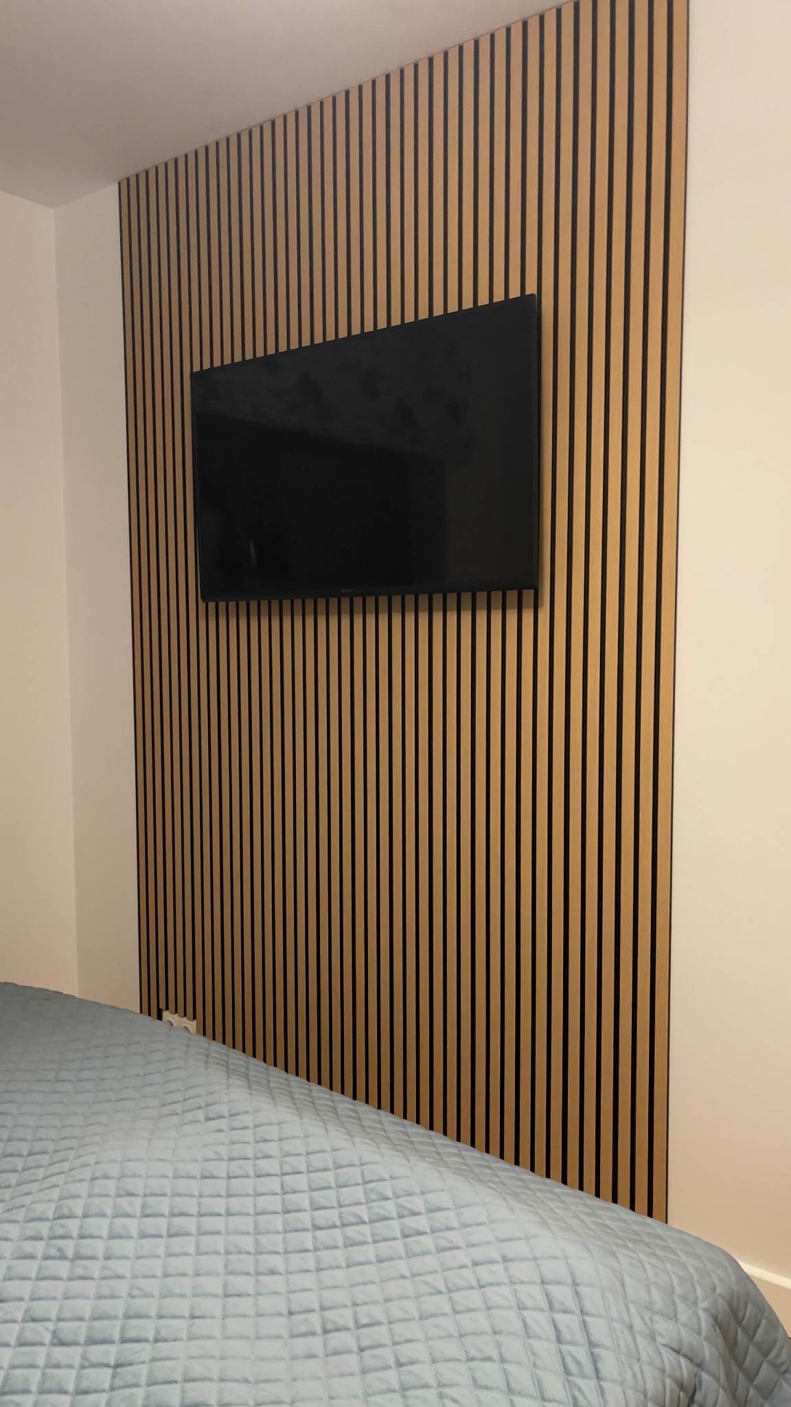 Telewizor zawieszony na ścianie z drewnianych paneli w sypialni, widoczny fragment łóżka z niebieską narzutą.