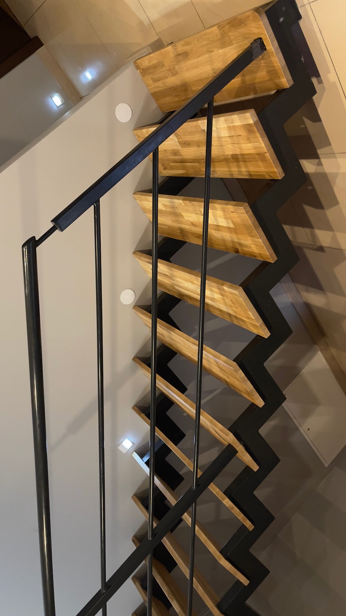 Nowoczesne schody drewniane z czarną metalową konstrukcją zygzakowatą i minimalistyczną balustradą, oświetlone punktowymi lampami w suficie.