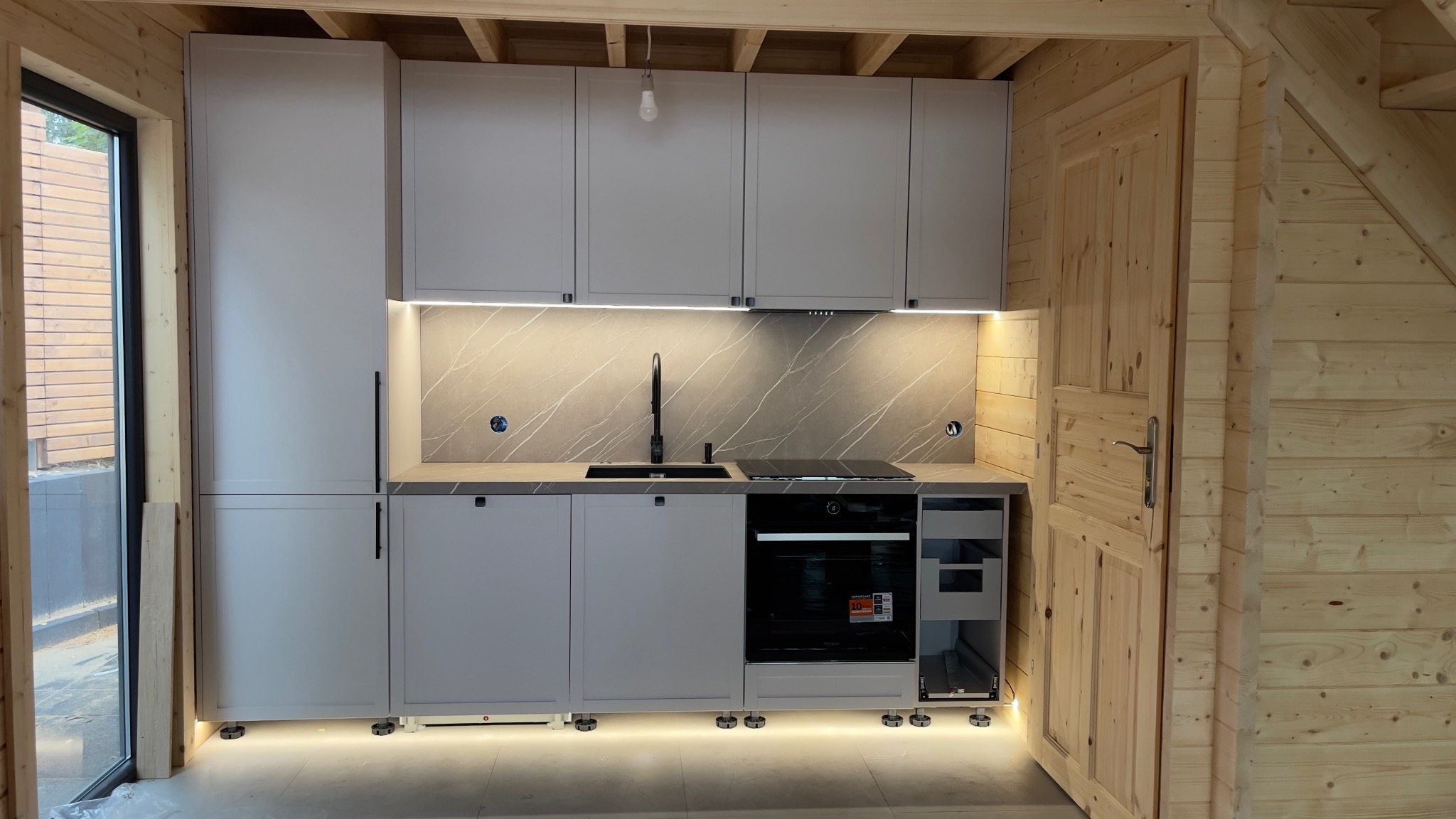 Jasna kuchnia w domku z bali: szare szafki z czarnymi uchwytami, blat imitujący marmur, czarna bateria i płyta indukcyjna, oświetlenie LED pod szafkami, widoczna drewniana konstrukcja ścian.