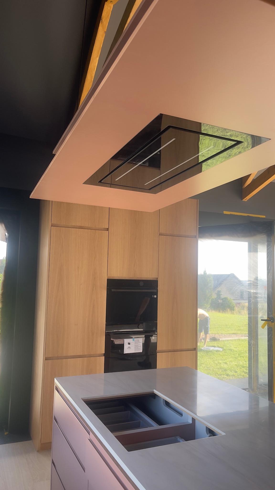 Nowoczesna kuchnia z jasnymi drewnianymi szafkami, różowym sufitem z wbudowanym okapem i wyspą kuchenną z widocznym wkładem na sztućce, widok na zewnątrz przez duże okno.