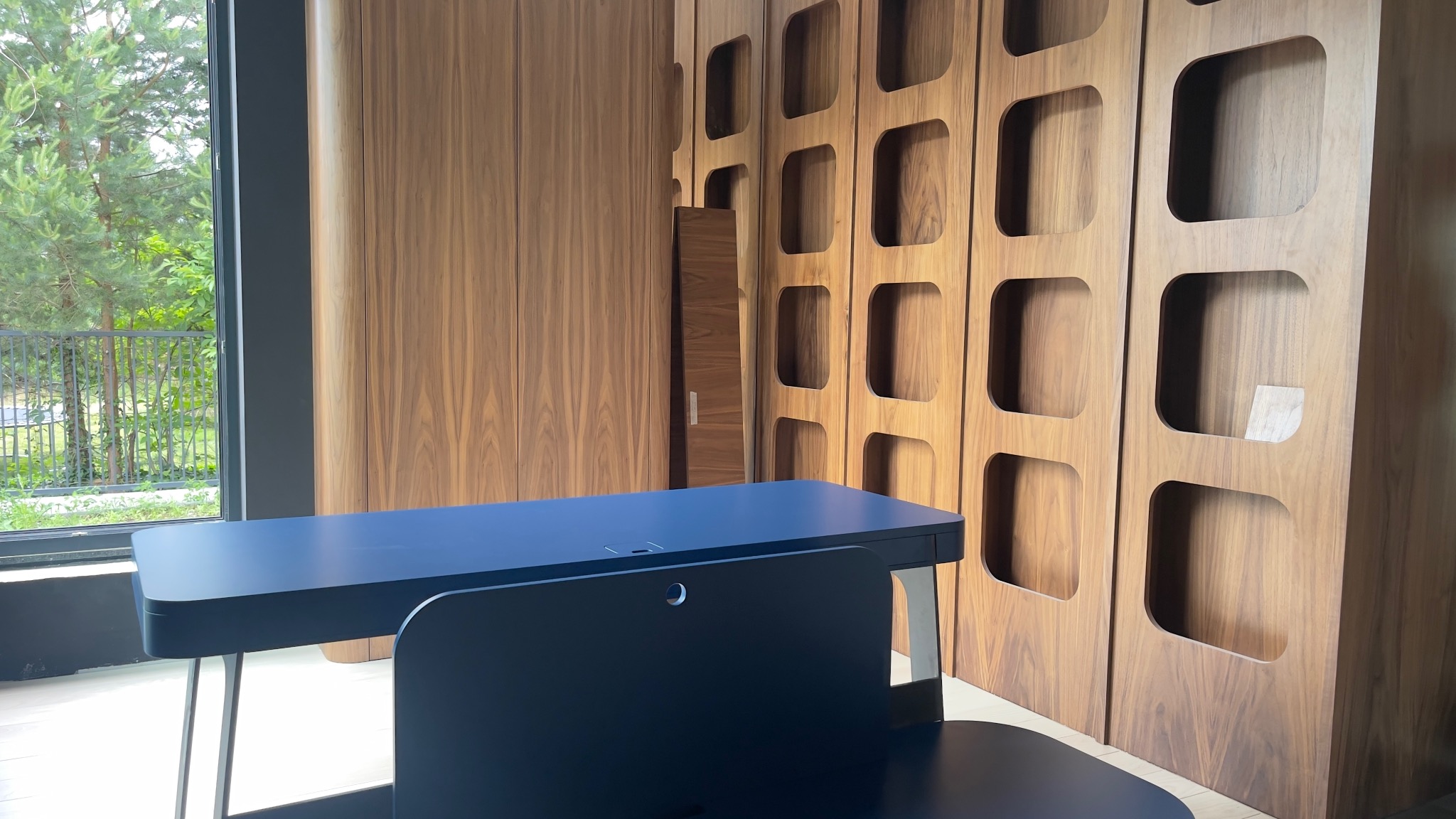 Nowoczesna szafa z drewna orzechowego z charakterystycznymi kwadratowymi wnękami, uzupełniona minimalistycznym biurkiem w kolorze granatowym na tle okna z widokiem na zieleń.