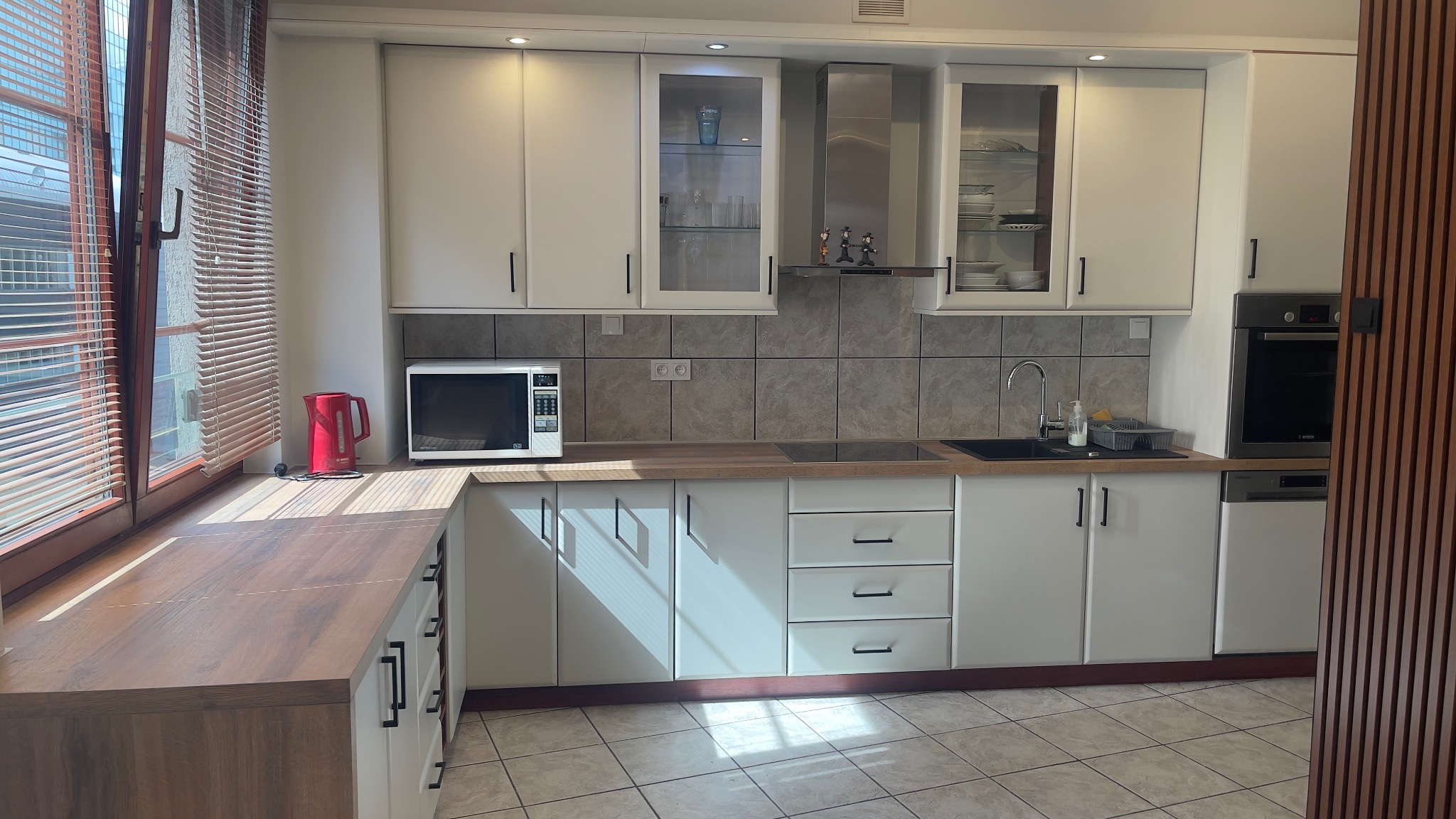 Jasna kuchnia z białymi szafkami, drewnianym blatem i szarymi płytkami, widok na okno z żaluzjami i czerwony czajnik.