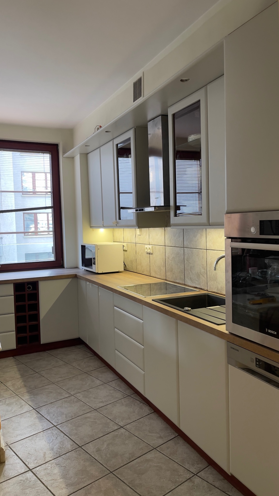 Jasna kuchnia z białymi szafkami, drewnianym blatem, zabudowaną kuchenką i zlewem, widok na okno z roletami i zabudowę na wino.