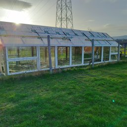 Leszek Kożdoń - Budowa szklarni z widocznymi panelami dachowymi w trakcie montażu, konstrukcja z białymi ramami okiennymi i drewnianymi słupkami, trawnik w pierwszym planie, słupy wysokiego napięcia w tle.