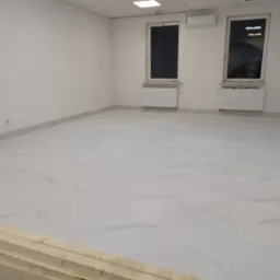 Wnętrze pustego pomieszczenia z nową, jasną podłogą z płytek imitujących marmur, dwa okna z grzejnikami pod spodem, klimatyzator na ścianie i stertą drewnianych desek w lewym dolnym rogu.