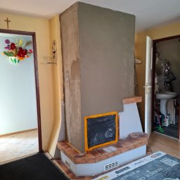 Leszek Kożdoń - Wnętrze z kominkiem w trakcie budowy/wykończenia. Widoczne ceglane elementy, surowy tynk i zabezpieczone folią miejsce na palenisko. W tle drabina i wejście do łazienki.