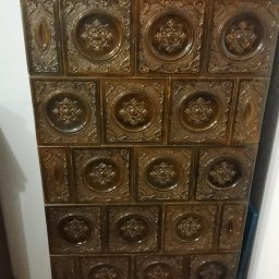 Leszek Kożdoń - Rustykalny piec kaflowy w odcieniach brązu, z dekoracyjnymi, wypukłymi wzorami na kaflach. Widoczne detale drzwiczek i ozdobne elementy na górze pieca.