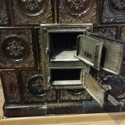 Leszek Kożdoń - Kafelkowy piec z otwartymi drzwiczkami paleniska, widoczne zdobienia na kaflach i narzędzie leżące na podłodze. Ujęcie z bliska.