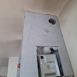 Leszek Kożdoń - Montaż obudowy kominka z szarej płyty, widoczne otwory wentylacyjne i podłączenie elektryczne. Ujęcie z dołu, sufit w tle. Etap prac wykończeniowych.