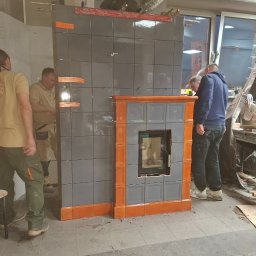Leszek Kożdoń - Wznoszenie kaflowego kominka z szarych i pomarańczowych płytek. Widoczne narzędzia i fragmenty instalacji. Prace w toku, kilku pracowników w tle.