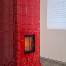 Leszek Kożdoń - Czerwony, kaflowy piec kominkowy z paleniskiem, zdobiony ornamentami, na tle ściany z wyświetlonymi zaletami użytkowania. Widoczny fragment podłogi z płytek.