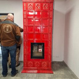 Leszek Kożdoń - Czerwony, kaflowy piec z szybą, zdobiony wzorami, w trakcie montażu przez dwóch mężczyzn. Widoczny fragment podłogi z szarych płytek.