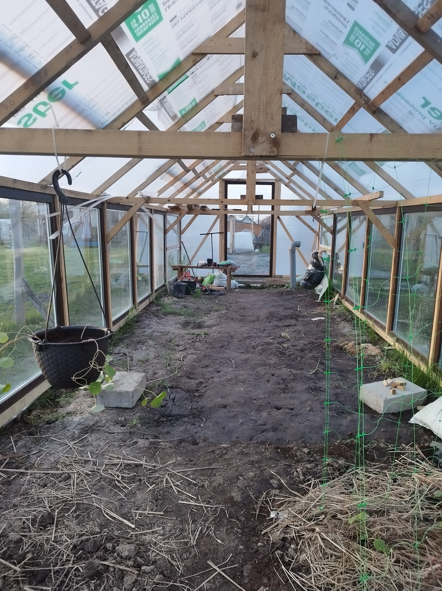 Wnętrze szklarni z drewnianą konstrukcją i przezroczystym dachem, widoczne donice, ziemia, siatka i betonowe bloczki, na zewnątrz widoczny ogród.