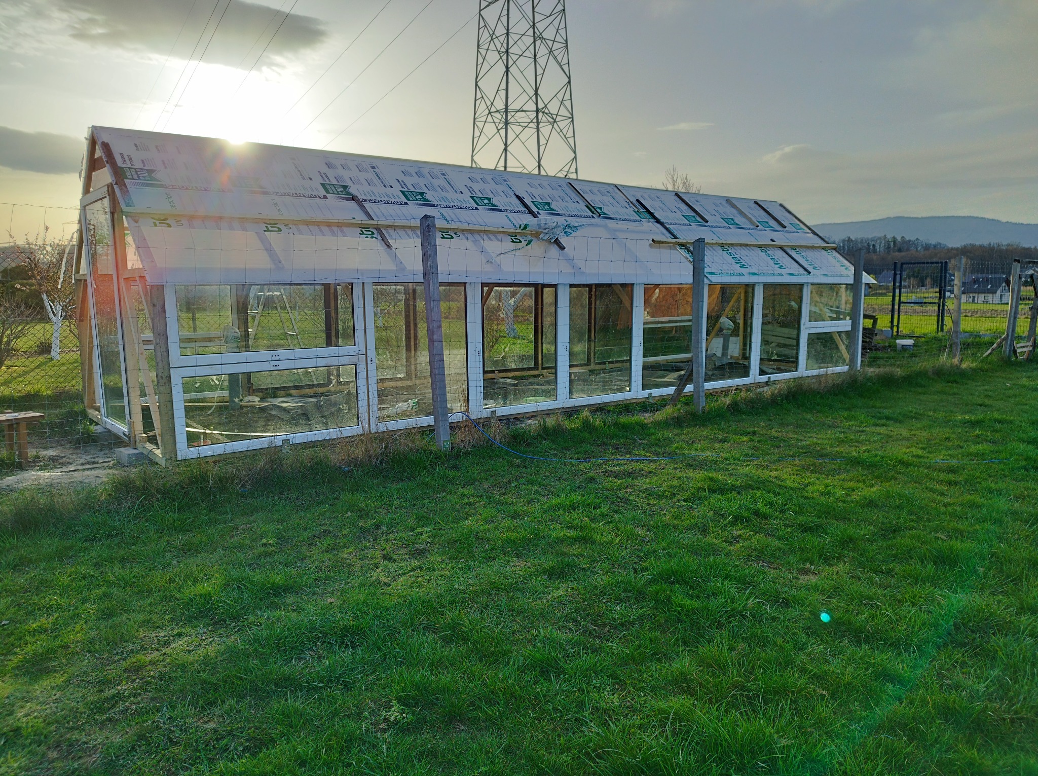 Budowa szklarni z widocznymi panelami dachowymi w trakcie montażu, konstrukcja z białymi ramami okiennymi i drewnianymi słupkami, trawnik w pierwszym planie, słupy wysokiego napięcia w tle.