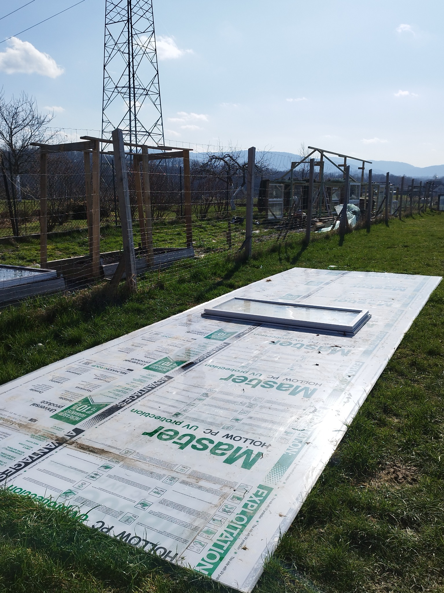 Płyta z poliwęglanu komorowego Master PC UV protection leżąca na trawie obok szkieletu konstrukcji szklarni z okien PCV i drewnianych belek na tle słupa wysokiego napięcia i gór.