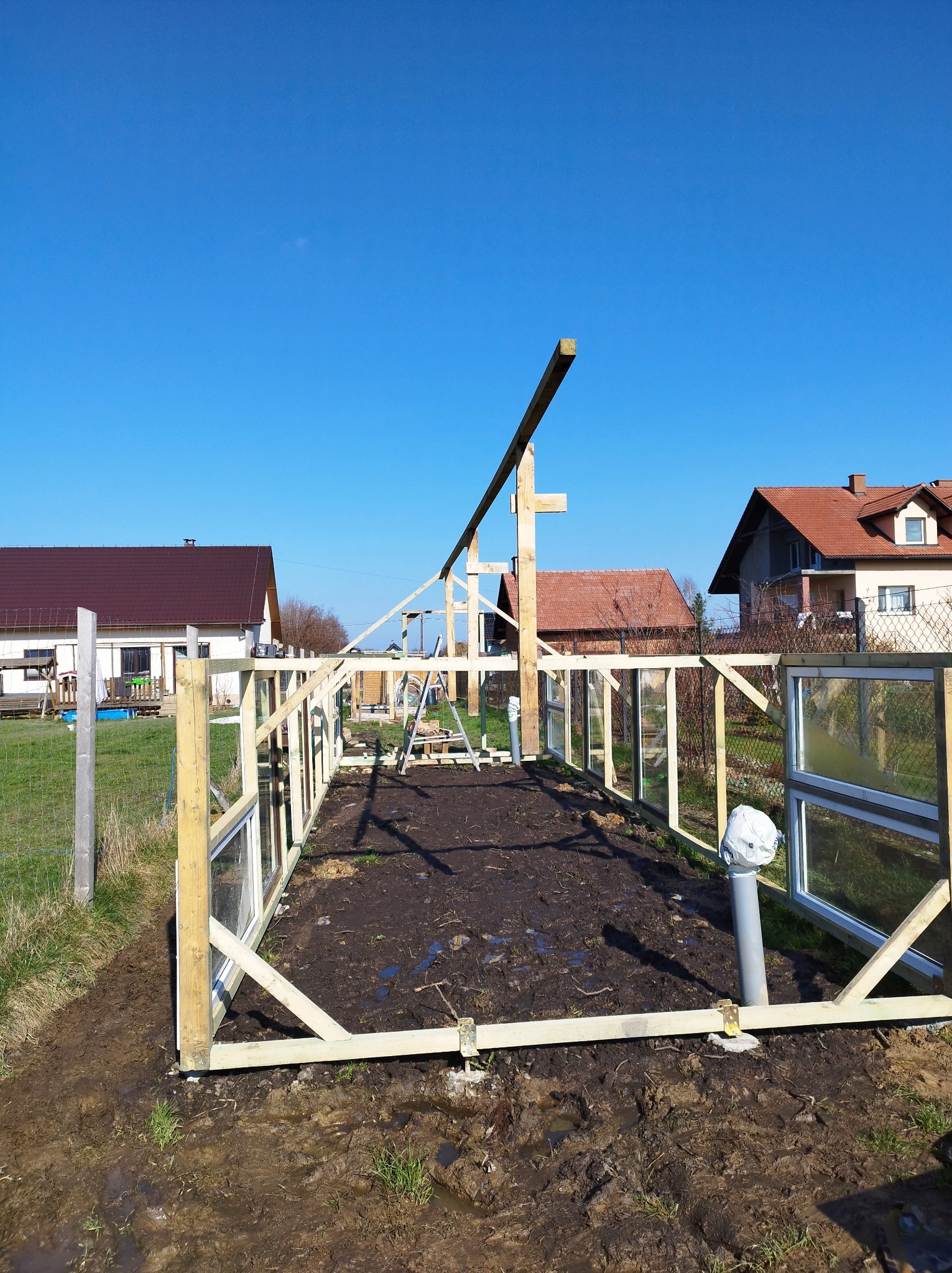 Konstrukcja szklarni z drewnianych ram i okien, widok z zewnątrz na tle domów i błękitnego nieba, widać etap budowy z częściowo ukończonym szkieletem.