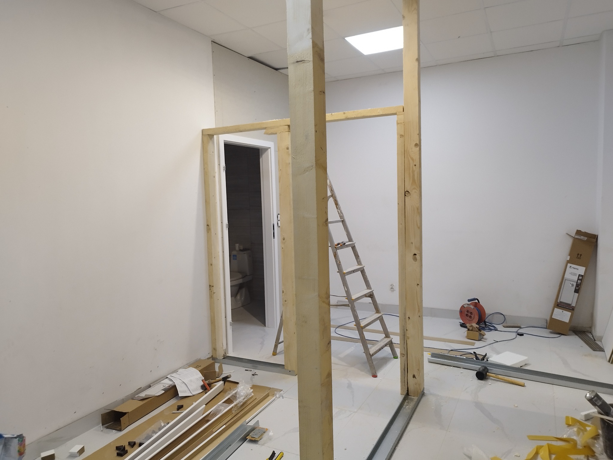 Wnętrze w trakcie budowy ścianki działowej z drewnianą ramą, widoczna drabina, profil metalowy podłogowy, narzędzia i materiały budowlane, w tle częściowo widoczna łazienka przez otwór drzwiowy.