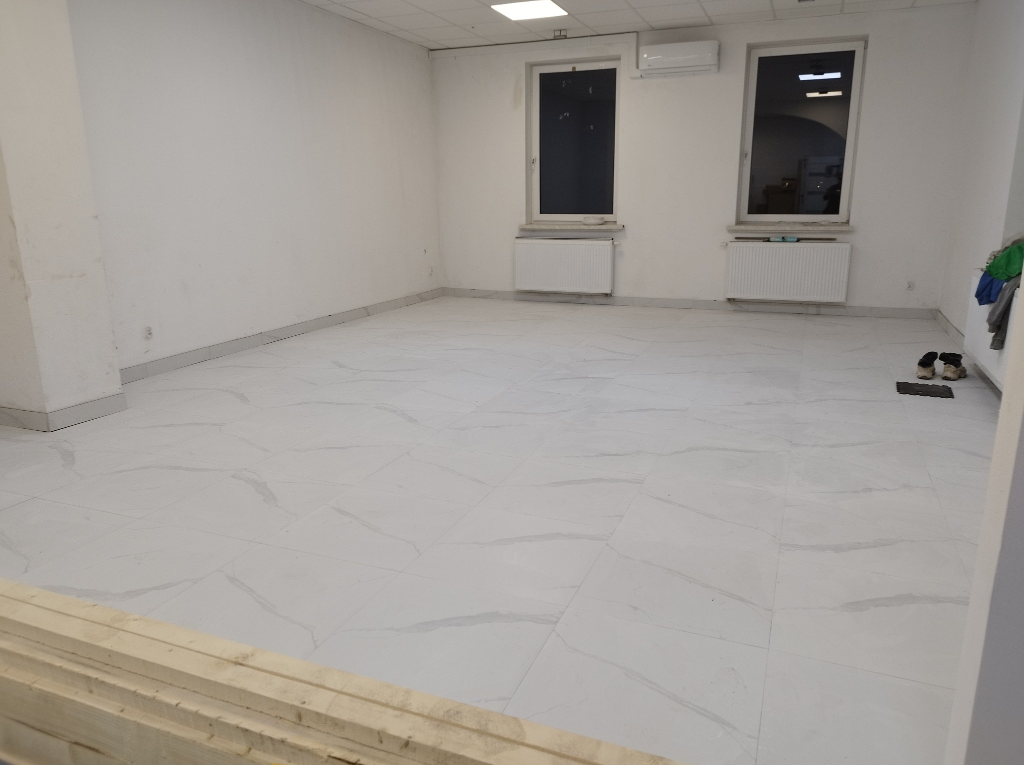 Wnętrze pustego pomieszczenia z nową, jasną podłogą z płytek imitujących marmur, dwa okna z grzejnikami pod spodem, klimatyzator na ścianie i stertą drewnianych desek w lewym dolnym rogu.