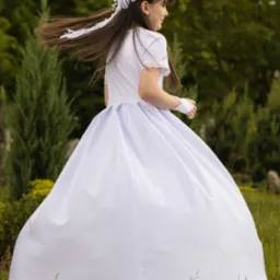 Dziewczynka w białej sukience komunijnej, z wiankiem na głowie, odwraca się, a jej włosy falują na wietrze na tle zieleni.