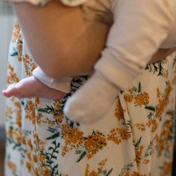 Arkadiusz Sęk Fotografia - Bliskie ujęcie bosej stopy niemowlęcia, delikatnie trzymanego przez dorosłą osobę w sukience z żółto-zielonym wzorem kwiatowym, z miękkim światłem.