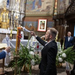 Arkadiusz Sęk Fotografia - Mężczyzna w czarnym garniturze zapala świecę od dużej, zdobionej paschałki w zabytkowym kościele. Obok, inni uczestnicy ceremonii z zapalonymi świecami i otwartą księgą, w tle ołtarz i kwiaty.