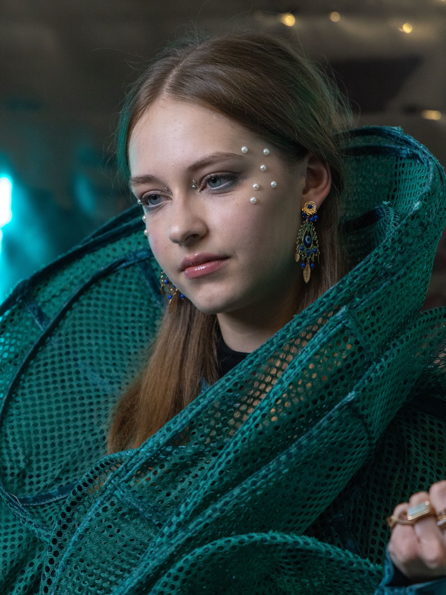 Portret młodej kobiety z perłami na twarzy, noszącej duże kolczyki z niebieskimi kamieniami i zieloną, ażurową szatę, lekko spoglądającej w dół.