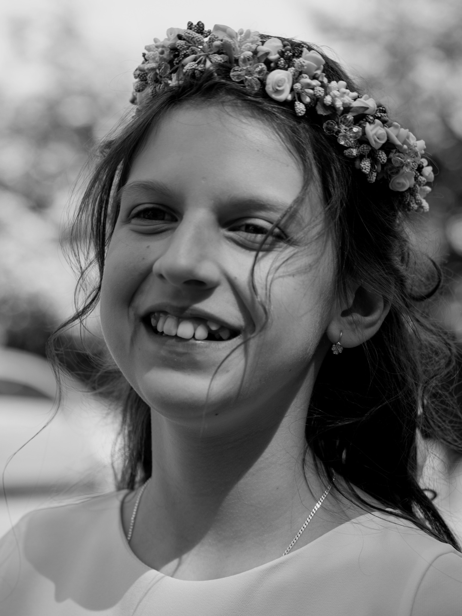 Portret uśmiechniętej dziewczynki w wianku z kwiatów i koralików, z delikatnym srebrnym łańcuszkiem i kolczykiem w kształcie kwiatka, uchwycony w czerni i bieli.