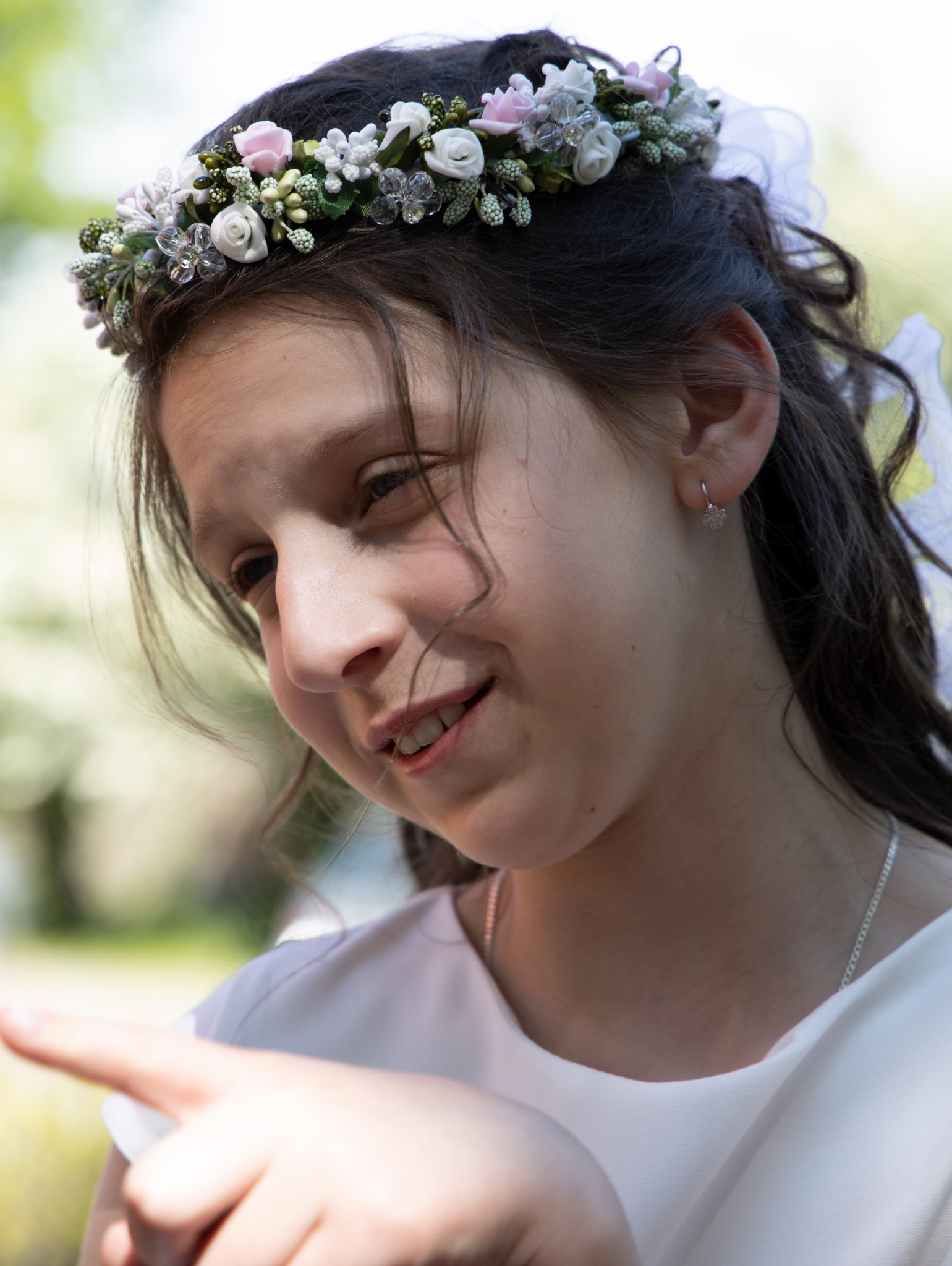 Portret dziewczynki z wiankiem z białych i różowych kwiatów na głowie, spoglądającej w dół i wskazującej palcem, z delikatnym uśmiechem i srebrnym naszyjnikiem.