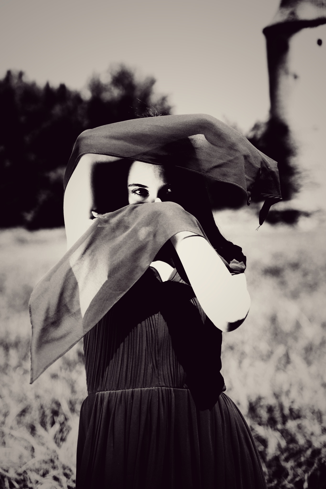 Czarno-białe zdjęcie kobiety w długiej sukni, z twarzą częściowo zasłoniętą materiałem, na tle polnego krajobrazu z wieżą w oddali, wykonane w Piotrków Trybunalski.