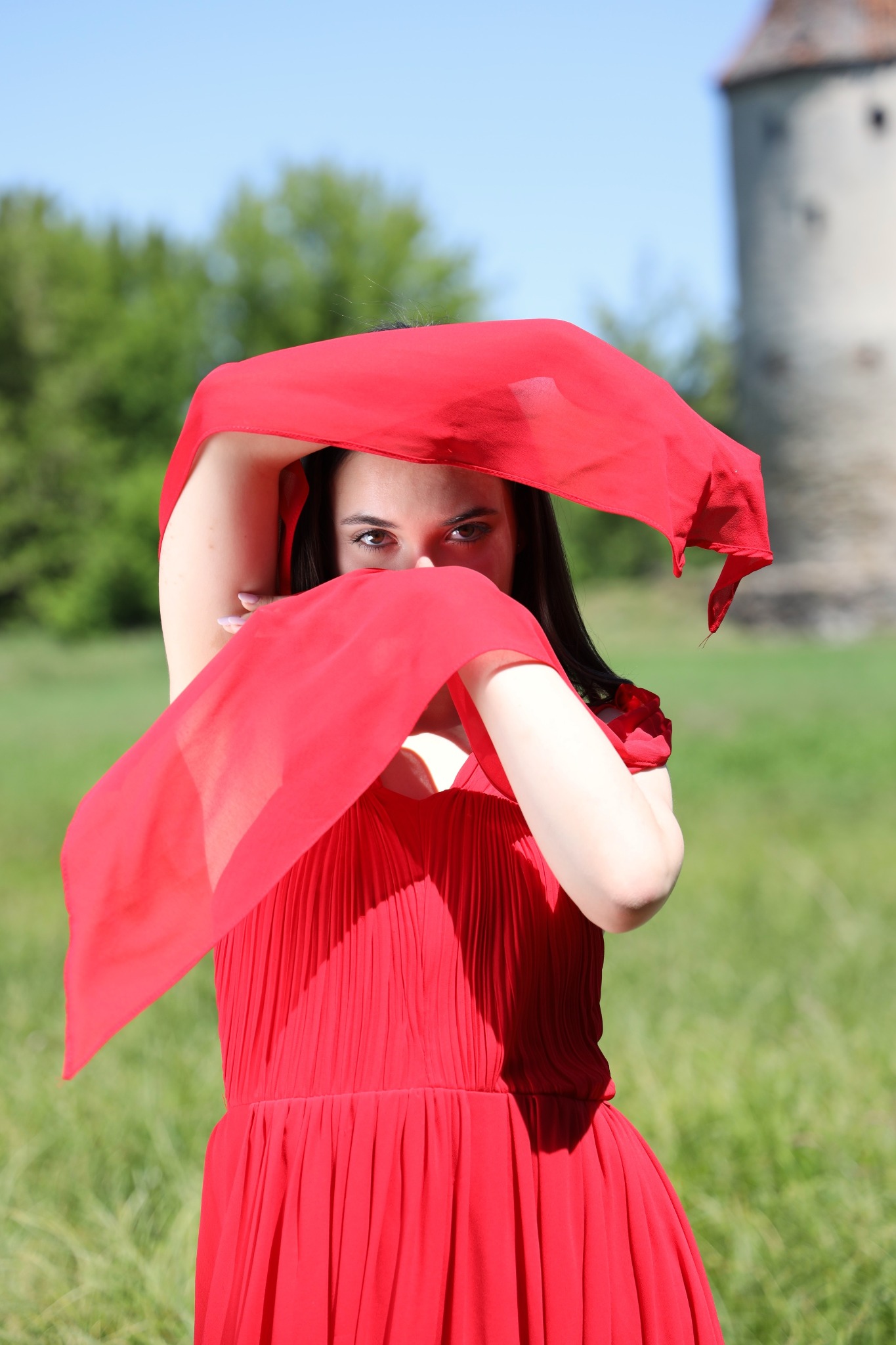 Kobieta w czerwonej sukience i z czerwoną chustą zakrywającą twarz, na tle zieleni i fragmentu starej wieży.