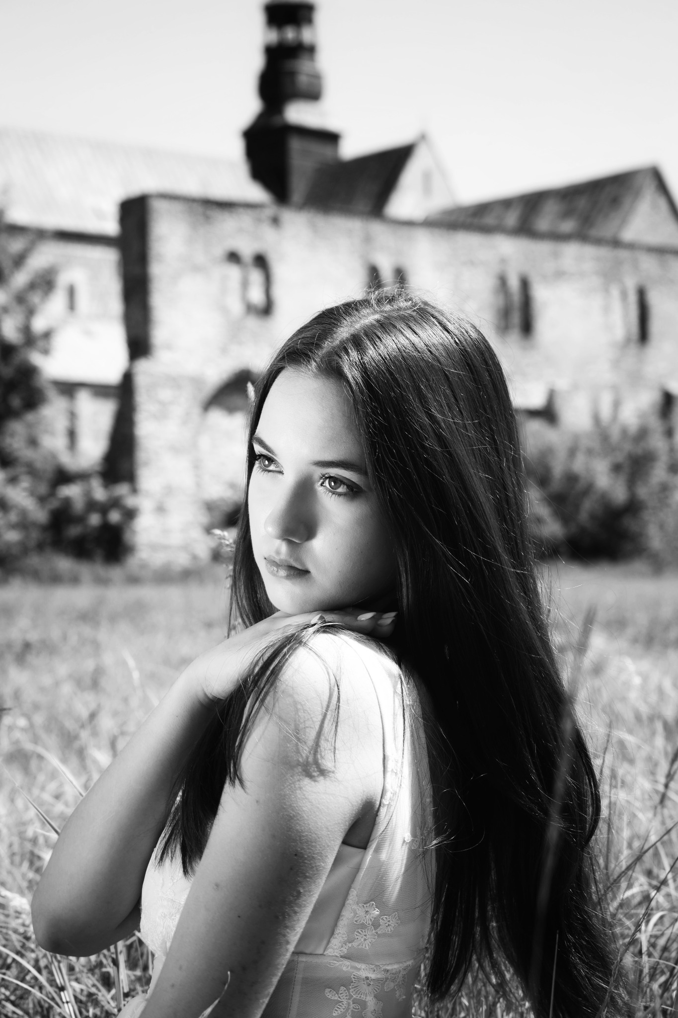 Portret młodej kobiety w sukience, opierającej brodę na dłoni, z zamyślonym spojrzeniem, na tle starego, kamiennego budynku w czerni i bieli.