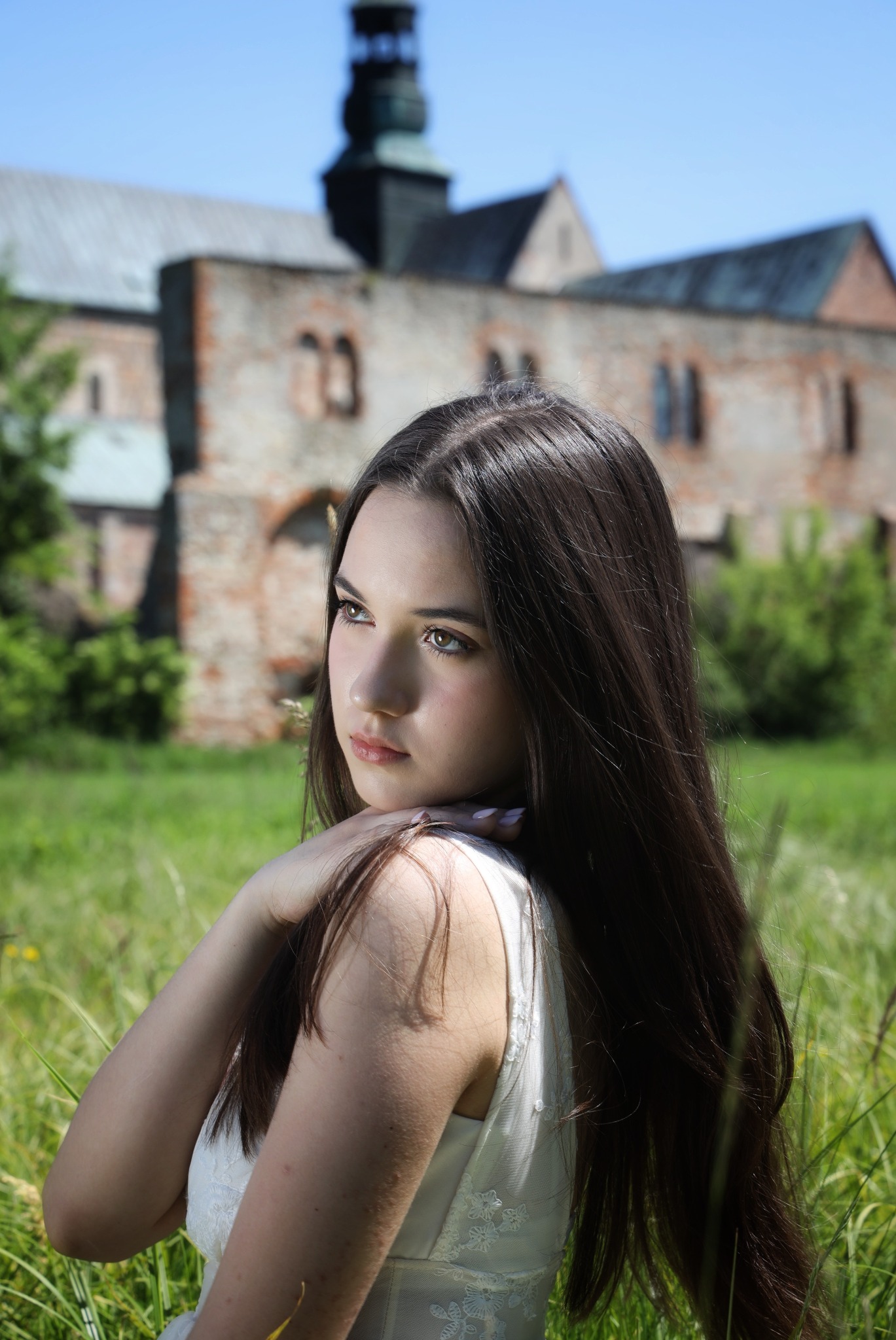 Portret młodej kobiety w białej sukience z koronkowymi detalami, pozującej na łące z wysoką trawą i zabytkowym budynkiem w tle.