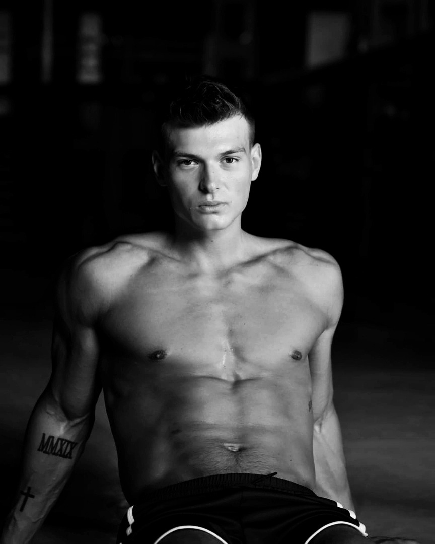 Portret czarno-biały muskularnego mężczyzny z tatuażem 'MMXIX' na ramieniu, spoglądającego w obiektyw na ciemnym tle.