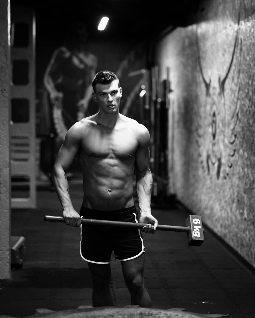 Czarno-białe zdjęcie muskularnego mężczyzny trzymającego młot o wadze 6 kg w siłowni z graffiti na ścianie w tle.