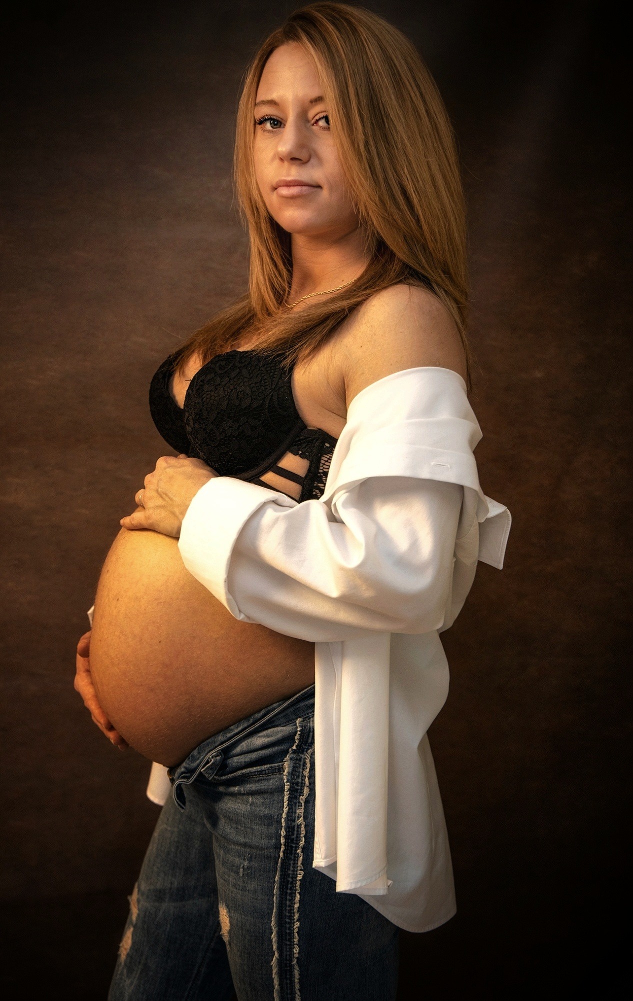 Portret kobiety w ciąży, w bieliźnie i rozpiętej białej koszuli, dżinsach, na brązowym tle studyjnym, z rękami na brzuchu.