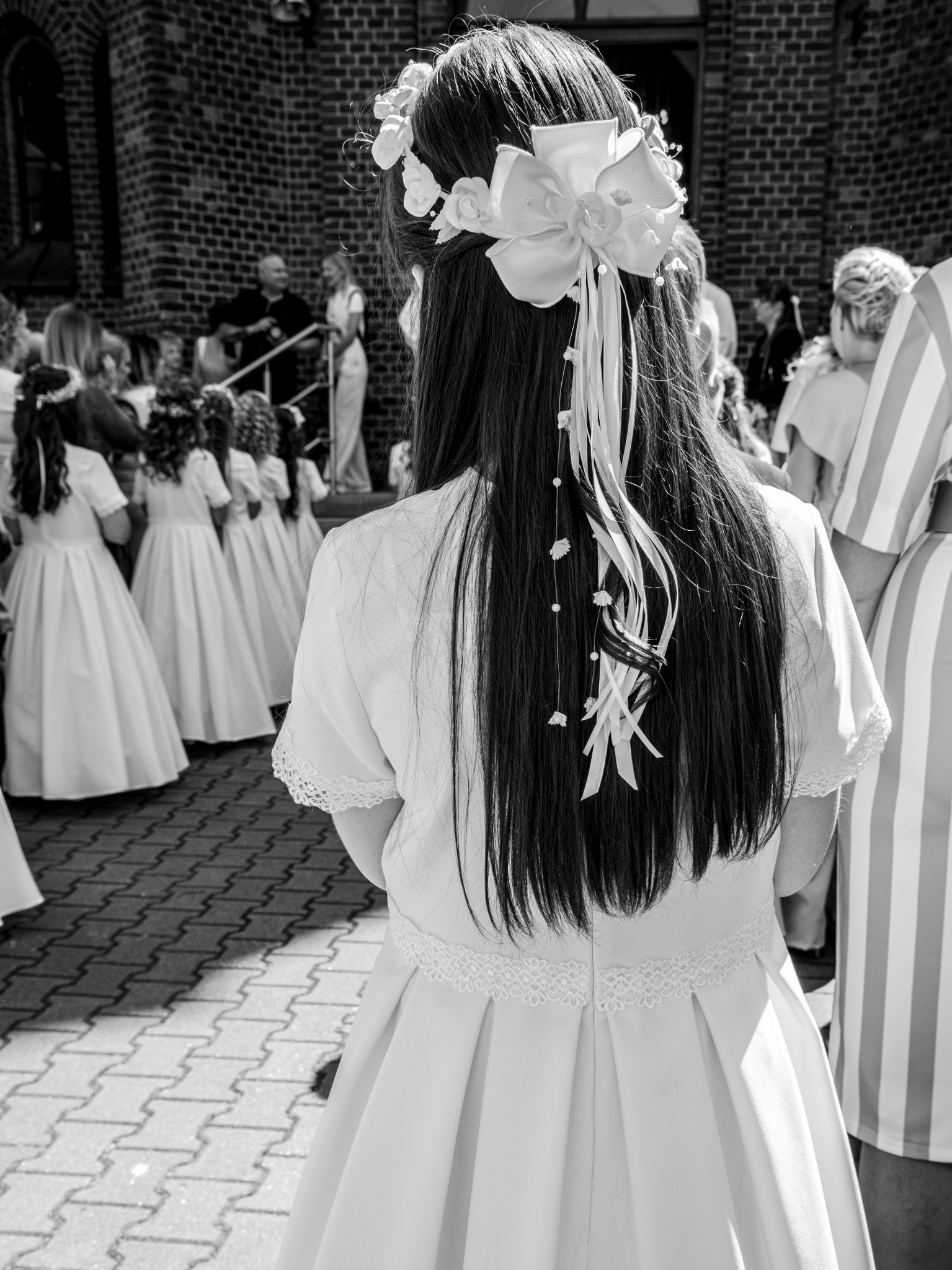 Czarno-białe zdjęcie z tyłu dziewczynki z długimi, ciemnymi włosami, w białej sukience z koronkowym wykończeniem i dużą białą kokardą z wstążkami we włosach, otoczonej innymi dziećmi w podobnych...