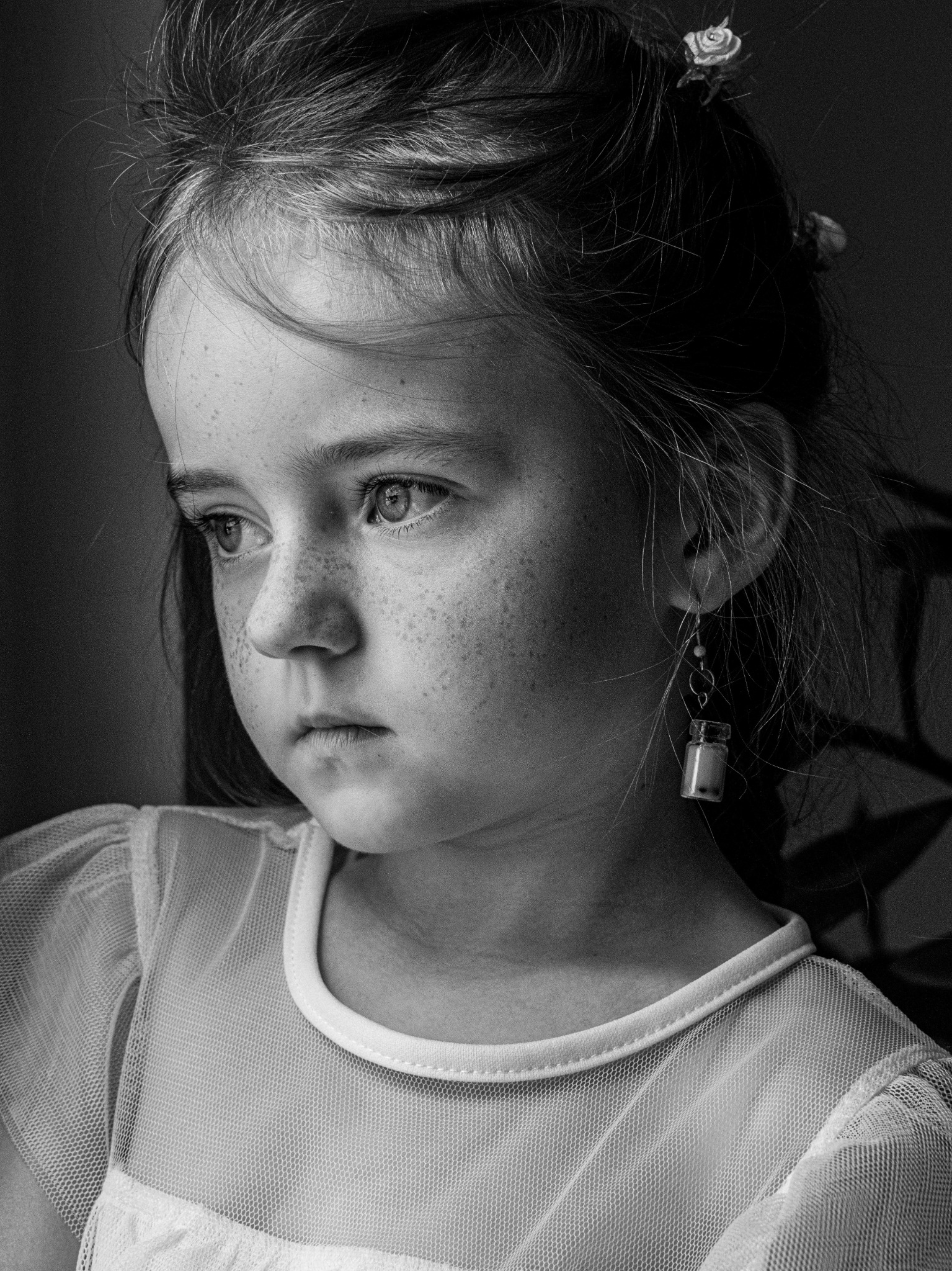 Portret dziewczynki z piegami, w sukience z tiulu, z wpiętymi we włosy drobnymi kwiatami i kolczykiem w kształcie małej buteleczki, czarno-biały kadr.