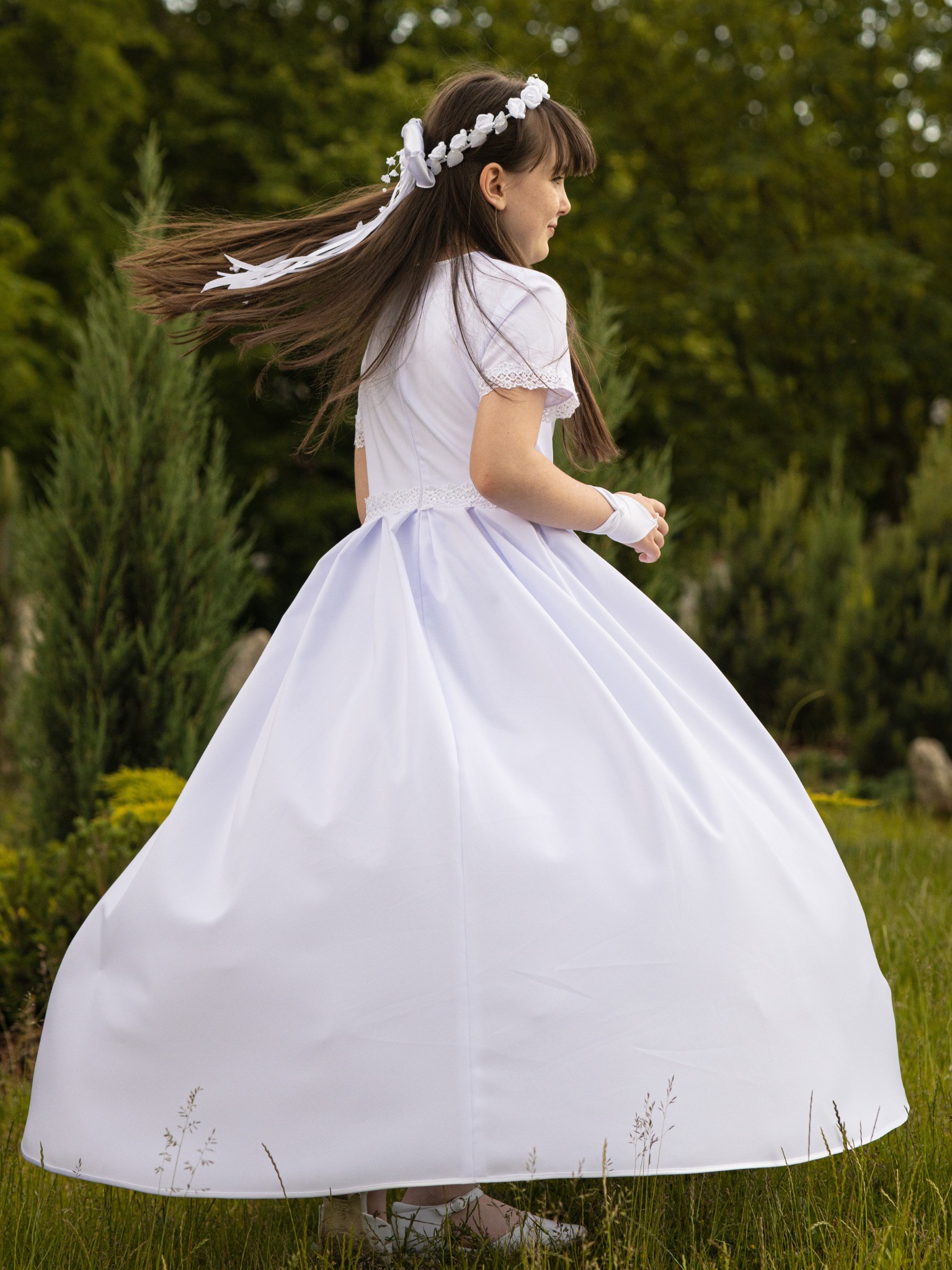Dziewczynka w białej sukience komunijnej, z wiankiem na głowie, odwraca się, a jej włosy falują na wietrze na tle zieleni.