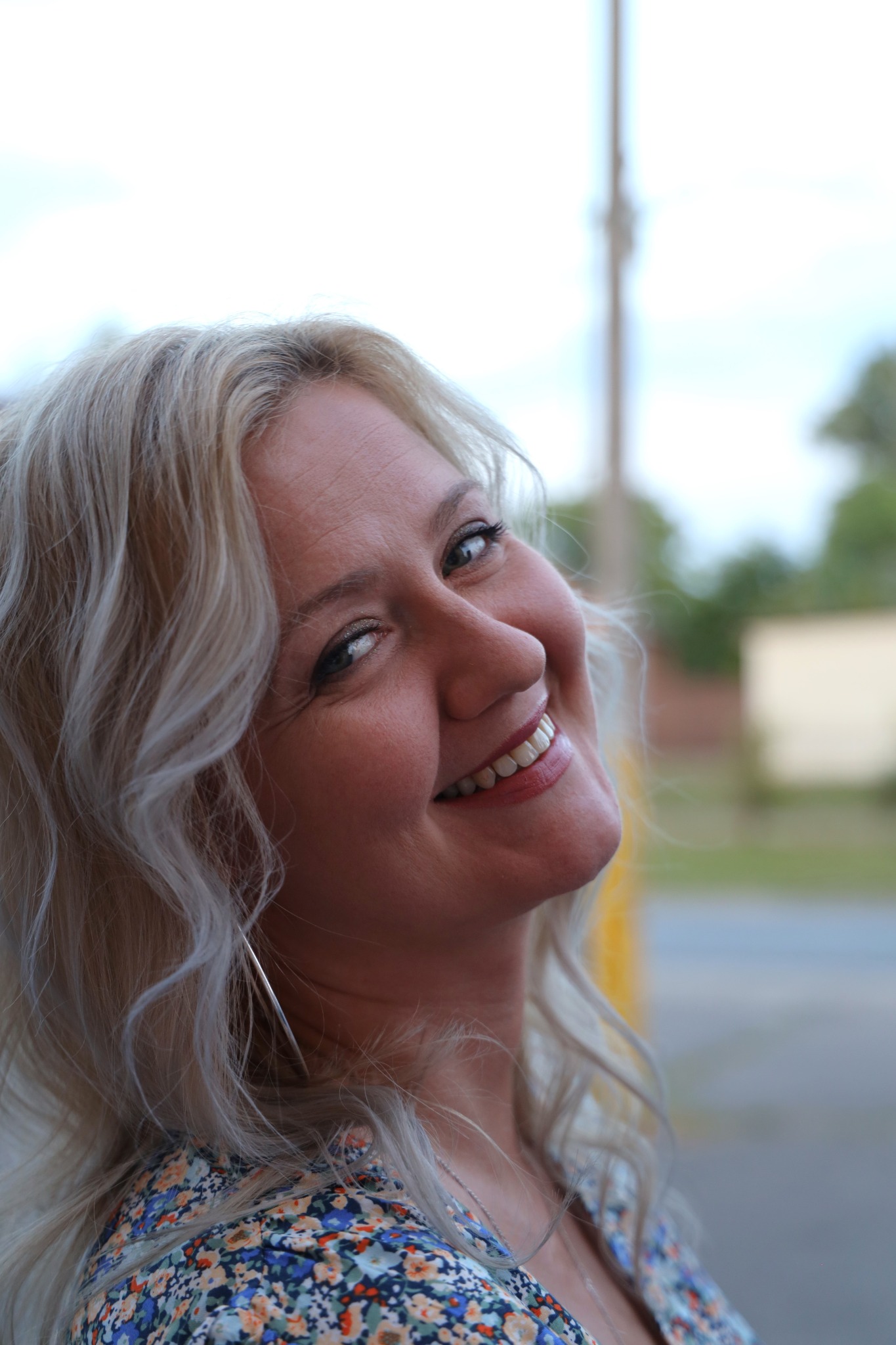 Uśmiechnięta kobieta z blond falowanymi włosami, srebrnymi kolczykami i kwiecistą bluzką, patrzy w górę w lewo.