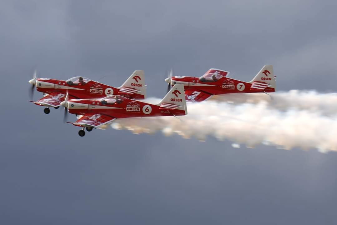 Trzy czerwono-białe samoloty akrobacyjne z logiem Orlen lecą w formacji, pozostawiając za sobą smugi dymu na tle pochmurnego nieba.