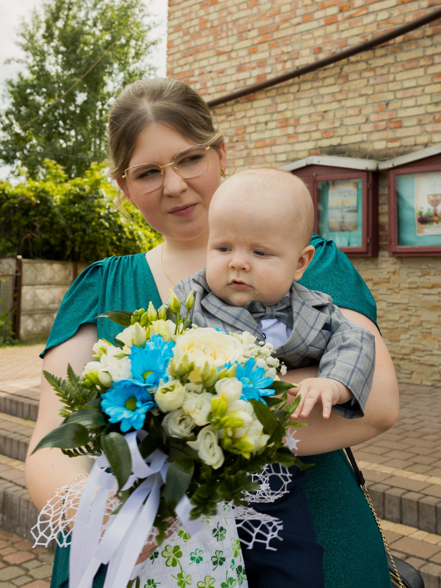 Kobieta w szmaragdowej sukience trzyma niemowlę w garniturze z bukietem białych i błękitnych kwiatów na tle ceglanej ściany i zieleni.