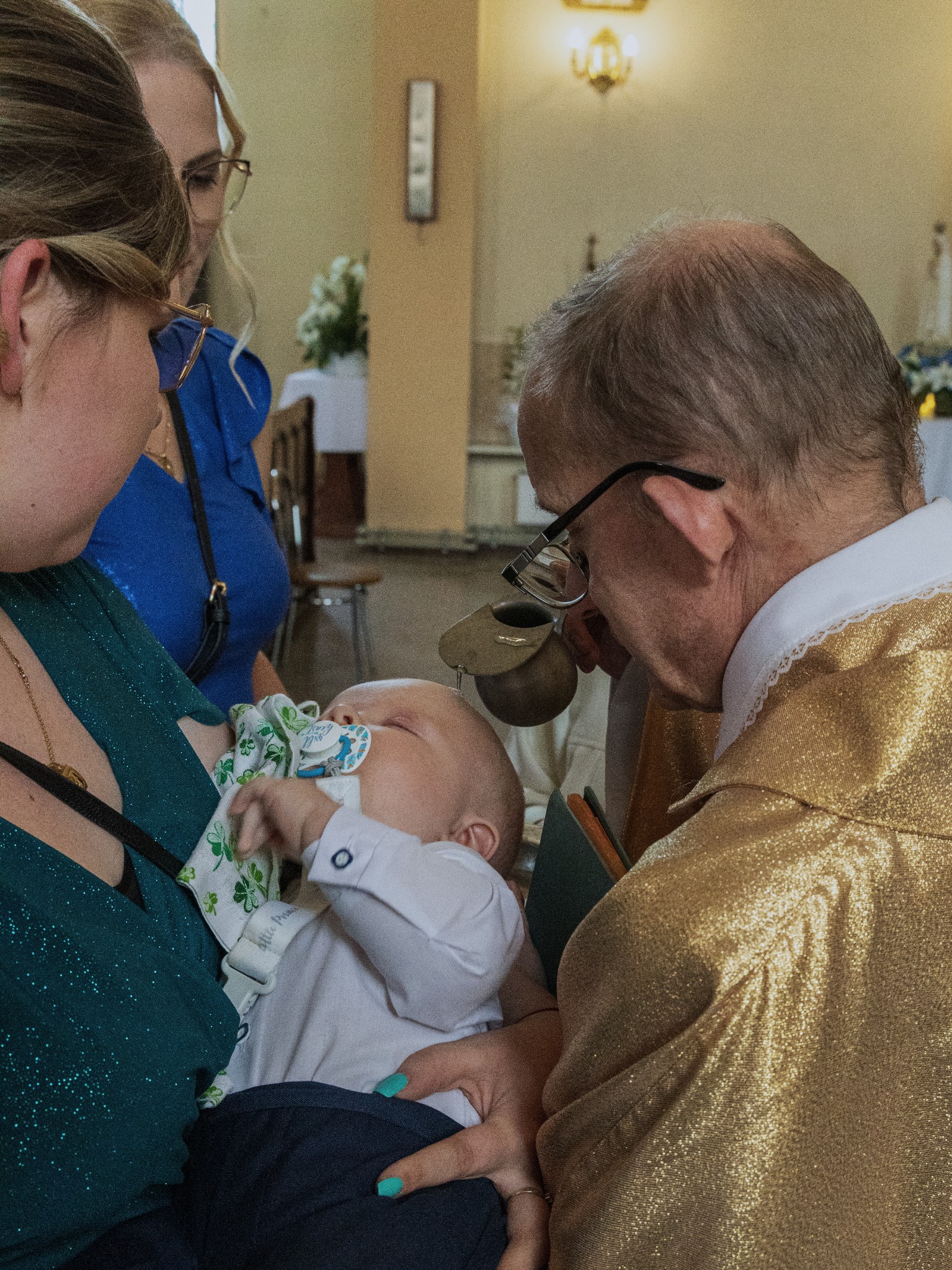 Ksiądz w złotej szacie udziela sakramentu chrztu śpiącemu niemowlęciu trzymanemu przez matkę w kościele. W tle widoczna kobieta w niebieskiej sukience.