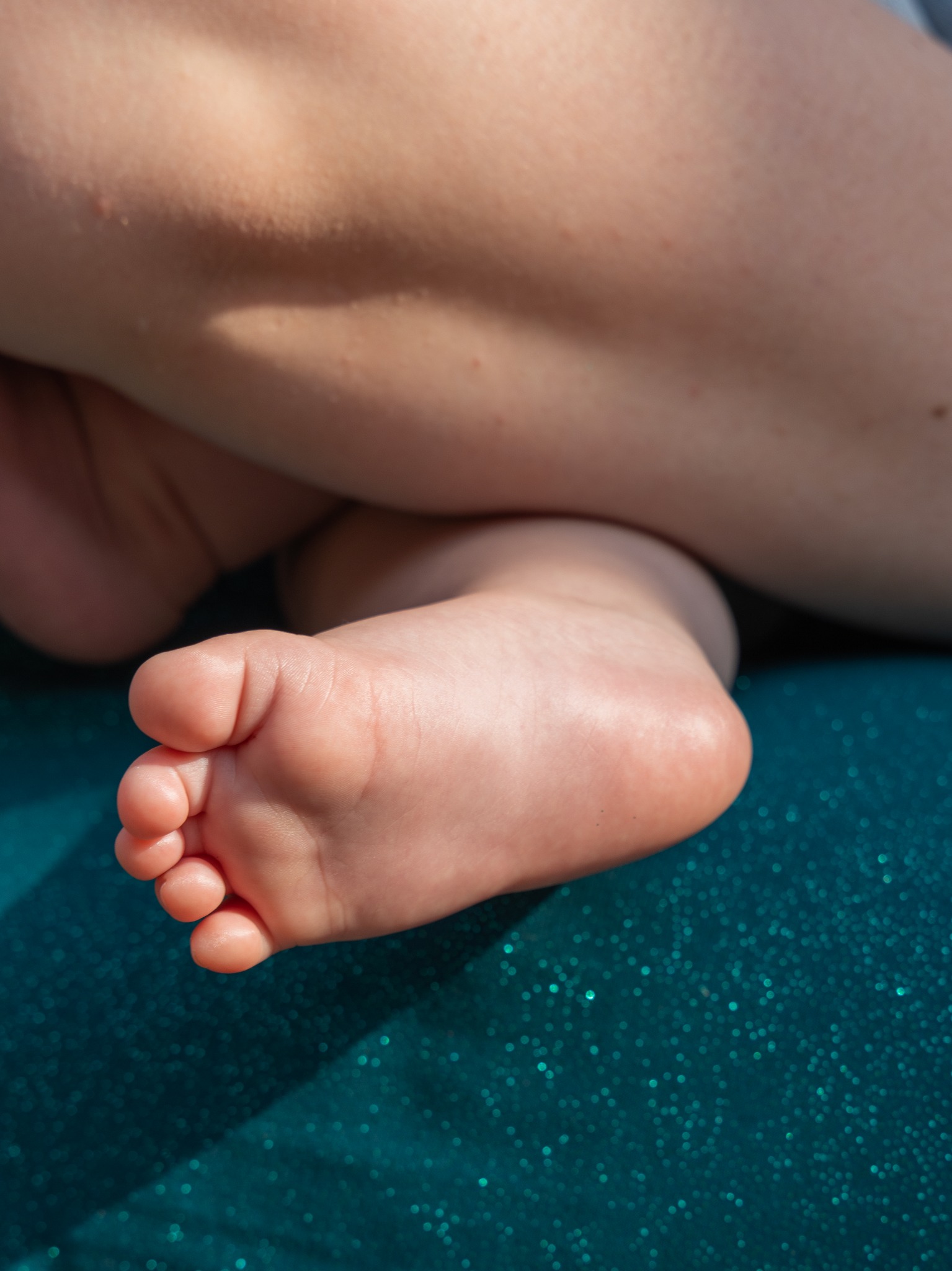 Mała stopa dziecka na turkusowym, połyskującym tle. Delikatne paluszki i fałdki skóry, ciepłe światło. Ujęcie z bliska, skupione na detalu stopy.