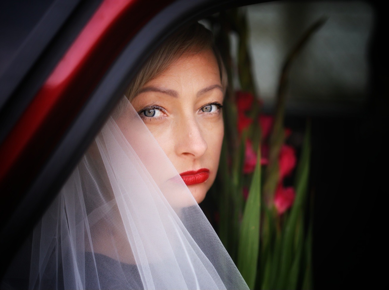 Panna młoda w samochodzie, częściowo zasłonięta woalką, z intensywnym makijażem ust i kwiatami w tle, spogląda w obiektyw zza ramy okna.
