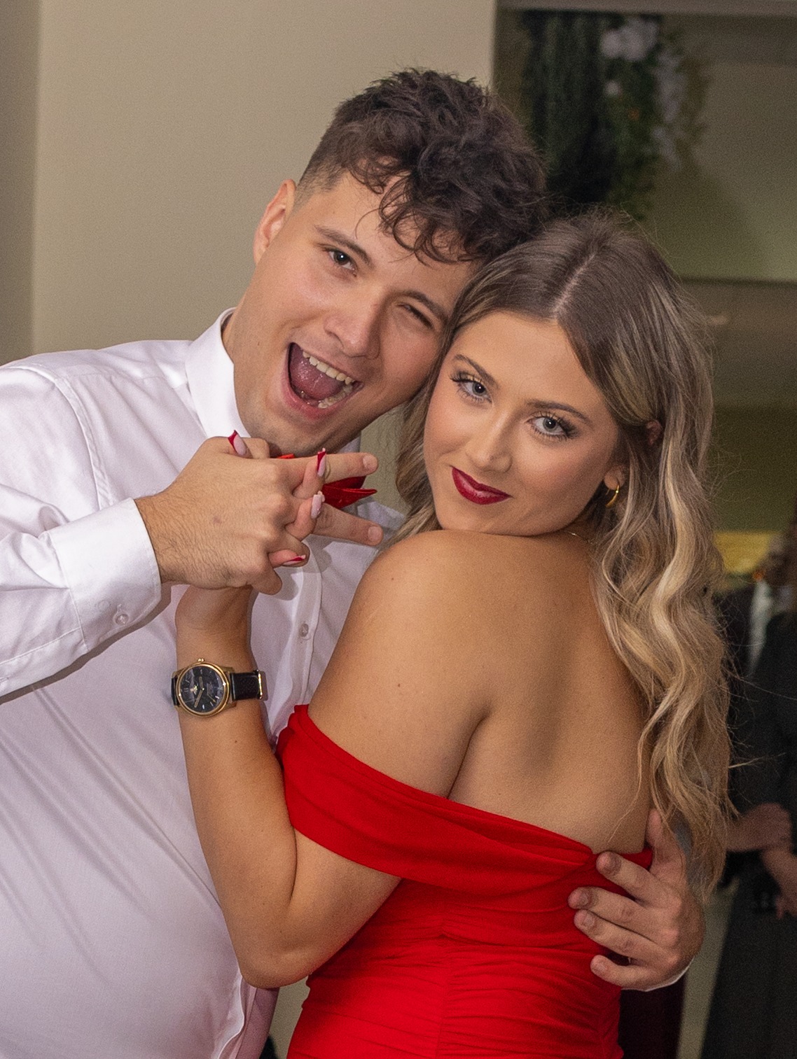 Uśmiechnięta para: mężczyzna mruga okiem, trzymając dłoń kobiety z czerwoną kokardką. Ona w czerwonej sukience, z makijażem i biżuterią. Zbliżenie, ciepłe światło.