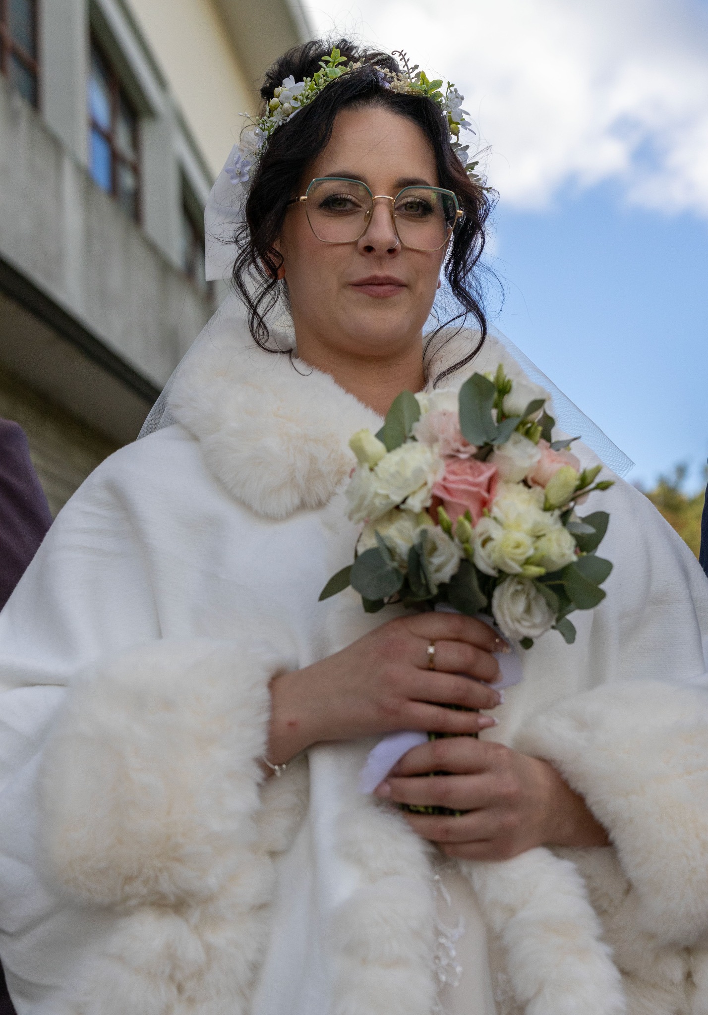 Panna młoda w białym futerku i okularach, trzymająca bukiet ślubny z różowych i białych kwiatów. W tle budynek i fragment nieba.